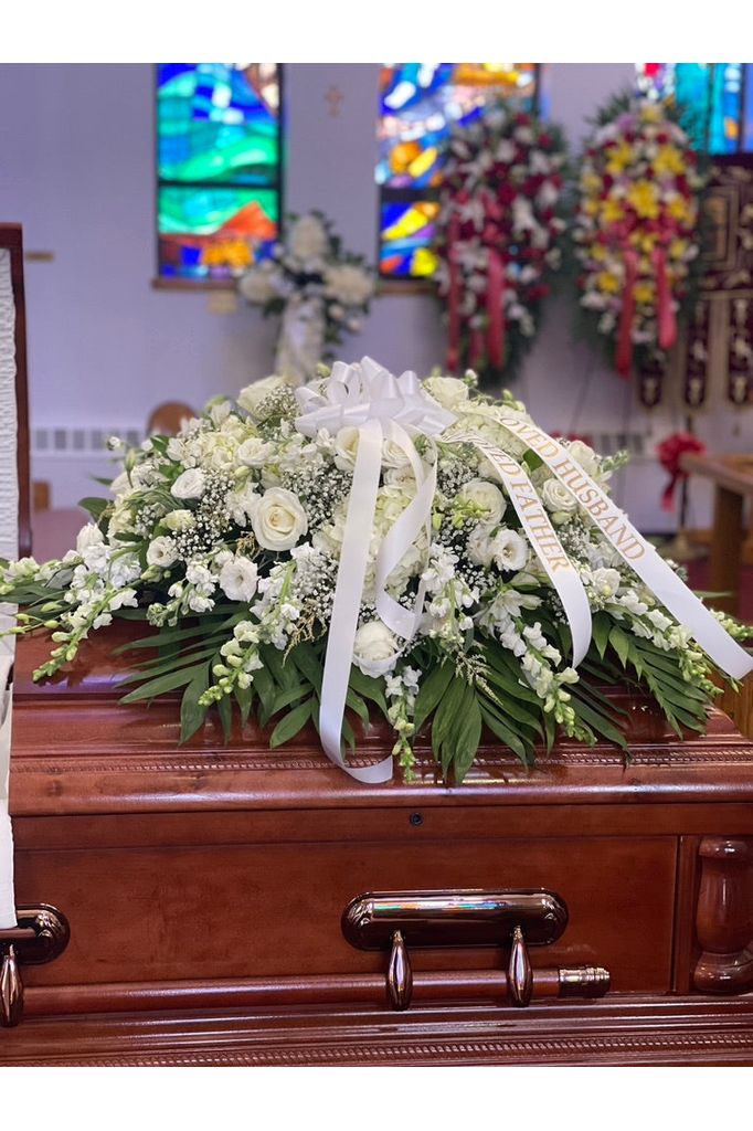 White funeral casket spray with roses, snapdragons, lisianthus, carnations, baby’s breath, and greenery placed on a wooden casket.
