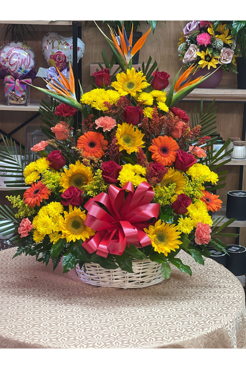 Bright sympathy floral basket with sunflowers, roses, carnations, gerbera daisies, and Birds of Paradise arranged in a white wicker basket.