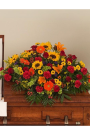 Bright multi-color casket spray with sunflowers, roses, lilies, gerberas, and carnations arranged on a wooden casket
