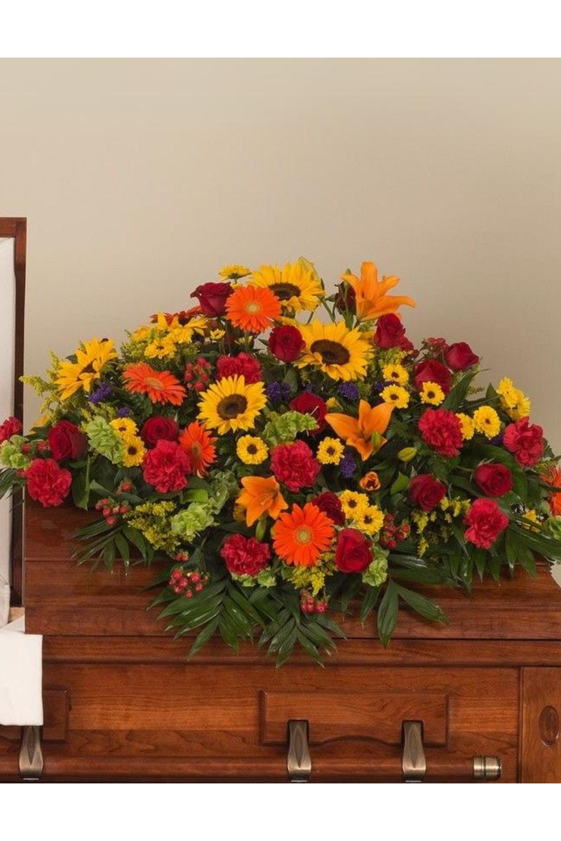 Bright multi-color casket spray with sunflowers, roses, lilies, gerberas, and carnations arranged on a wooden casket
