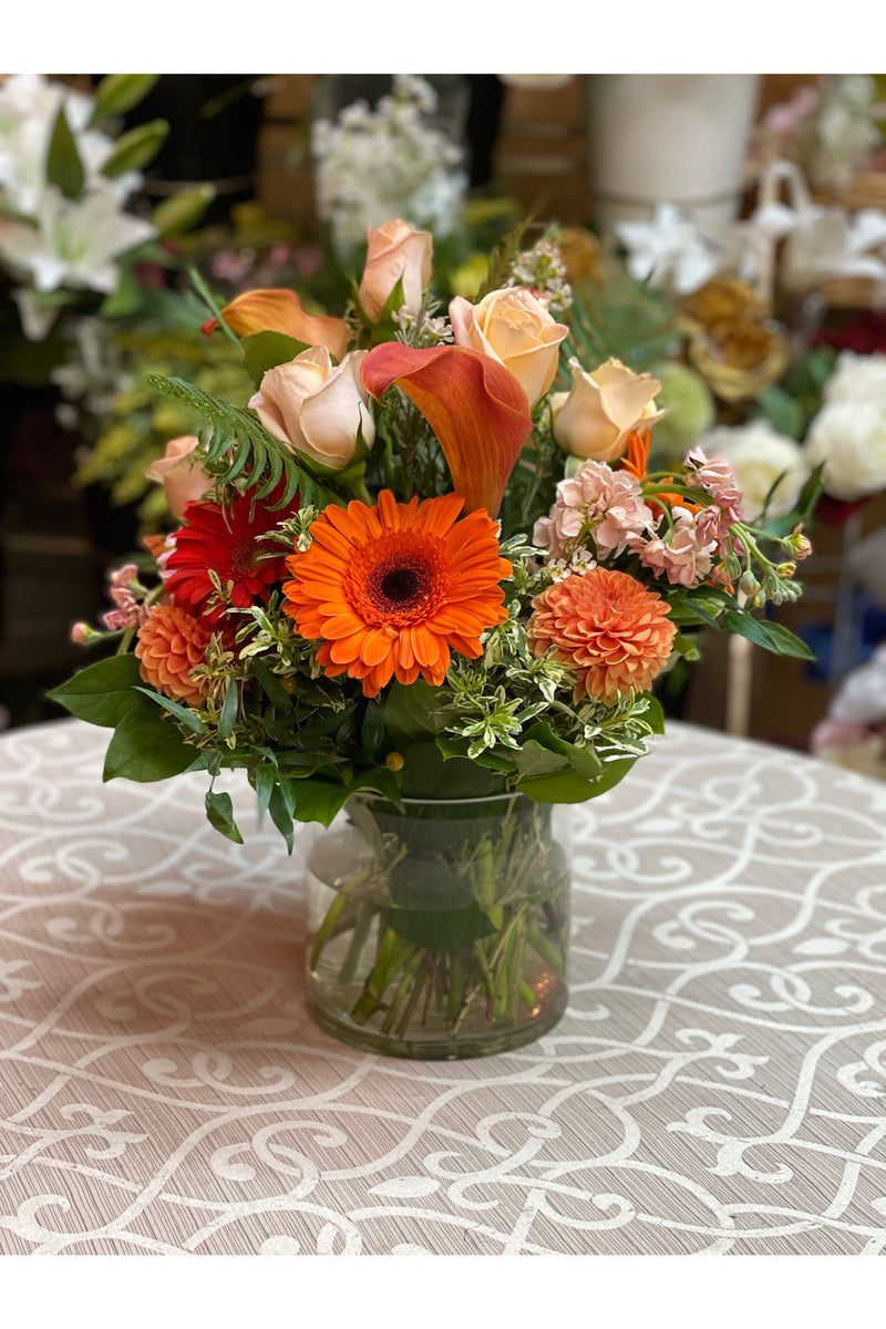 Vibrant orange flower arrangement with gerbera daisies, roses, calla lilies, and dahlias in a clear glass vase.