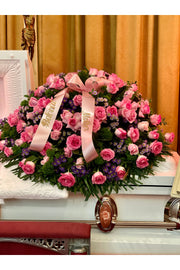 Pink rose funeral casket spray with lush greenery, purple asters, and pastel ribbons displayed on a casket.