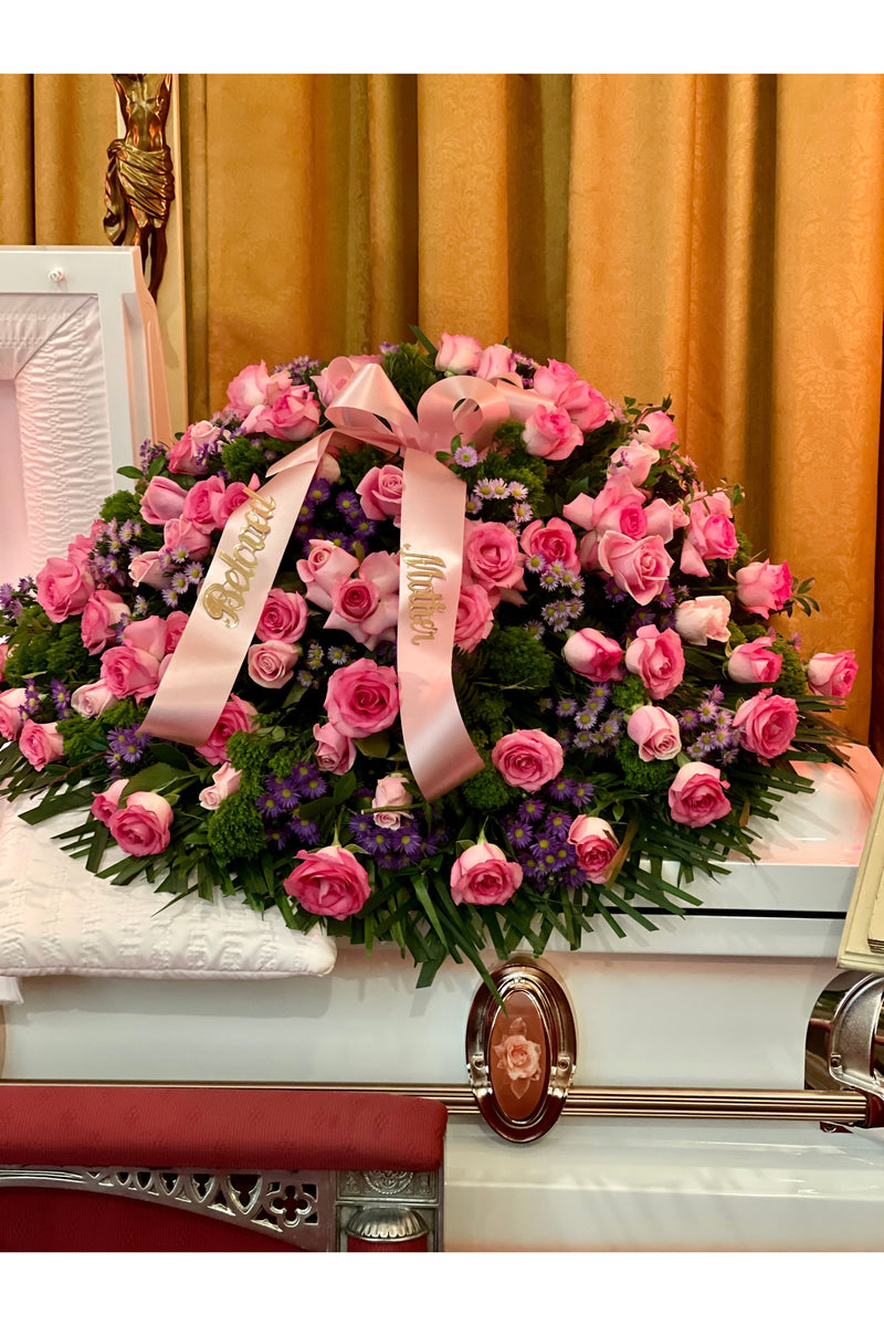 Pink rose funeral casket spray with lush greenery, purple asters, and pastel ribbons displayed on a casket.