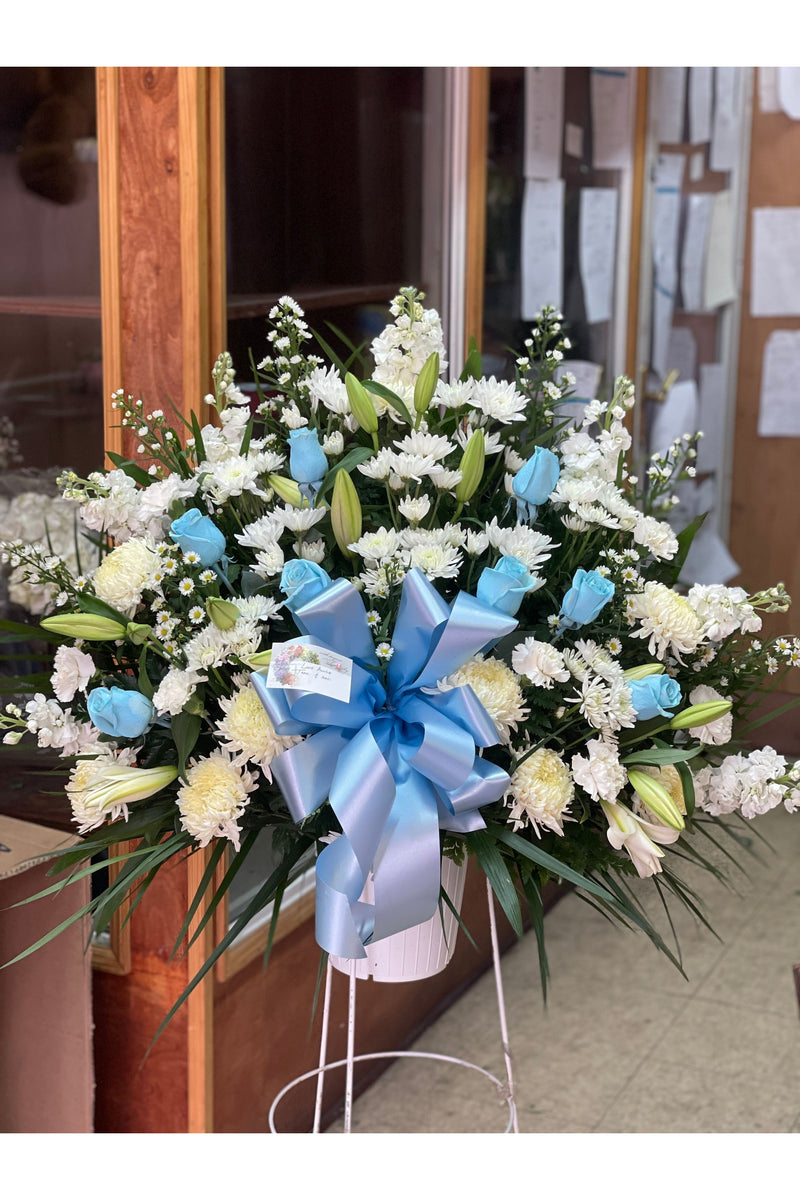 White and blue sympathy flower basket with lilies, blue roses, mums, and a large blue bow.