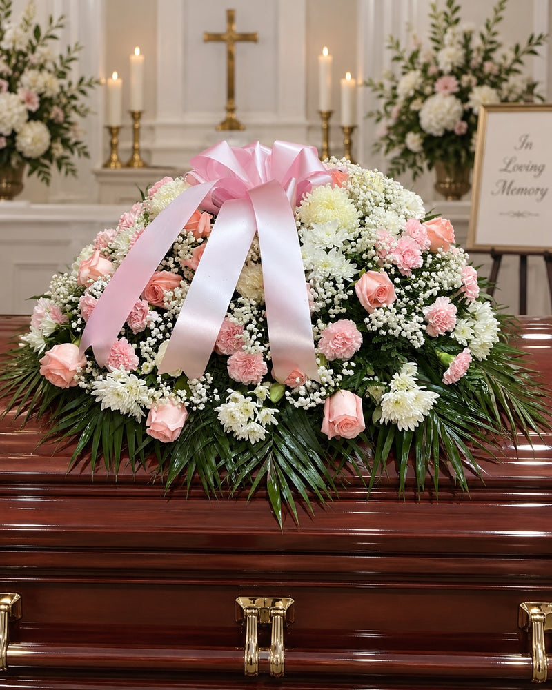 Pink and white funeral casket spray with roses, chrysanthemums, carnations and blush ribbon on a mahogany casket