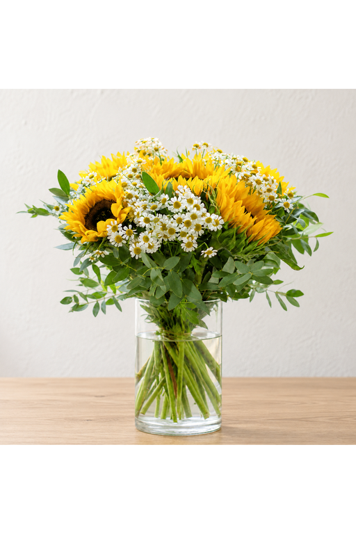 Yellow sunflowers and small white daisies with green foliage in a clear cylindrical glass vase
