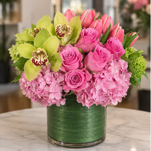 Strawberry Shortcake flower arrangement with pink roses, tulips, hydrangea and green orchids in a leaf-wrapped cylinder vase.
