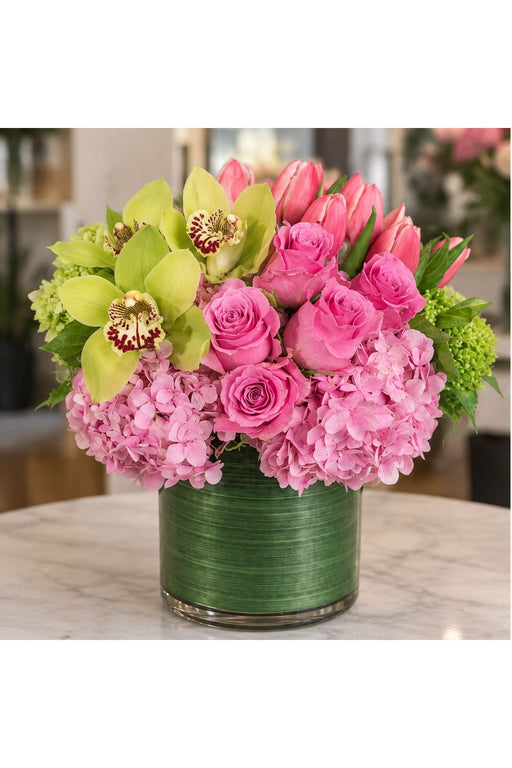 Strawberry Shortcake flower arrangement with pink roses, tulips, hydrangea and green orchids in a leaf-wrapped cylinder vase.