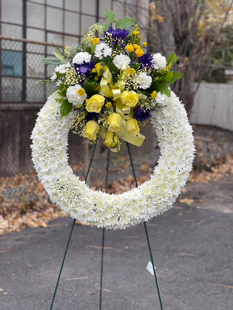 White sympathy wreath with yellow roses and purple accent flowers on easel