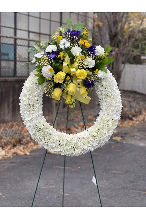 White sympathy wreath with yellow roses and purple accent flowers on easel