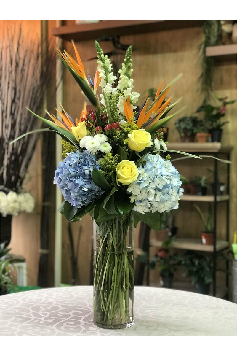 Tall flower arrangement with Birds of Paradise, blue hydrangeas, yellow roses, snapdragons, and greenery in a glass vase – New Jersey florist.