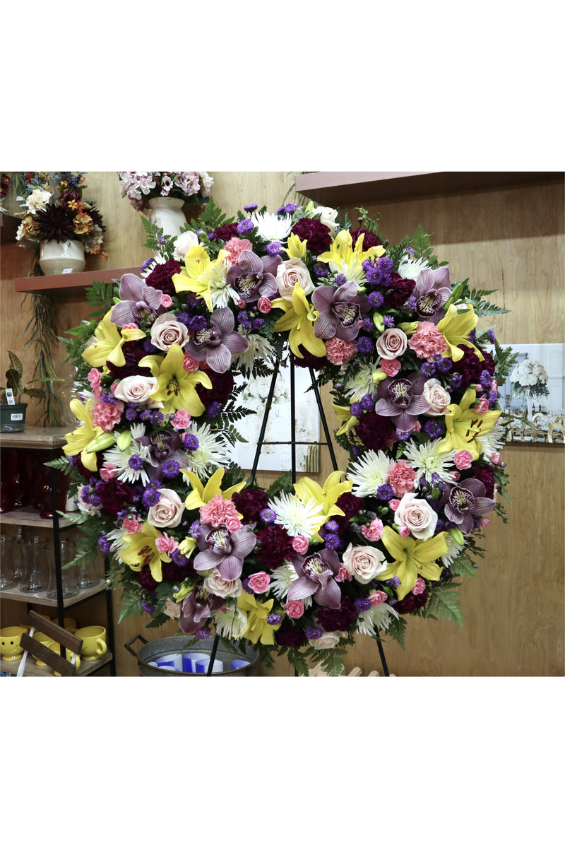 Purple and yellow funeral wreath with lilies, roses, orchids, and mixed blooms on a standing easel