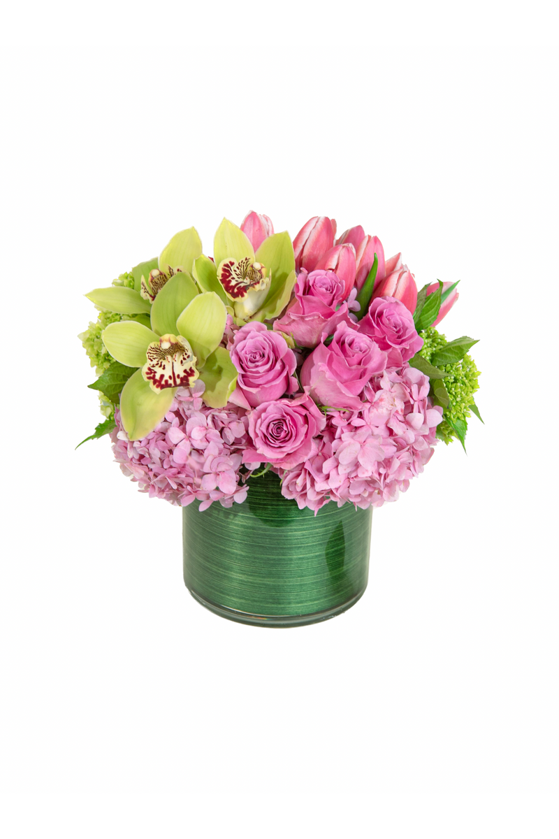 Strawberry Shortcake arrangement with pink roses, tulips, hydrangea, and green orchids in a green glass vase.