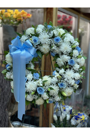 White and blue funeral wreath with white mums, lilies, blue roses, and large blue bow – standing sympathy floral tribute.