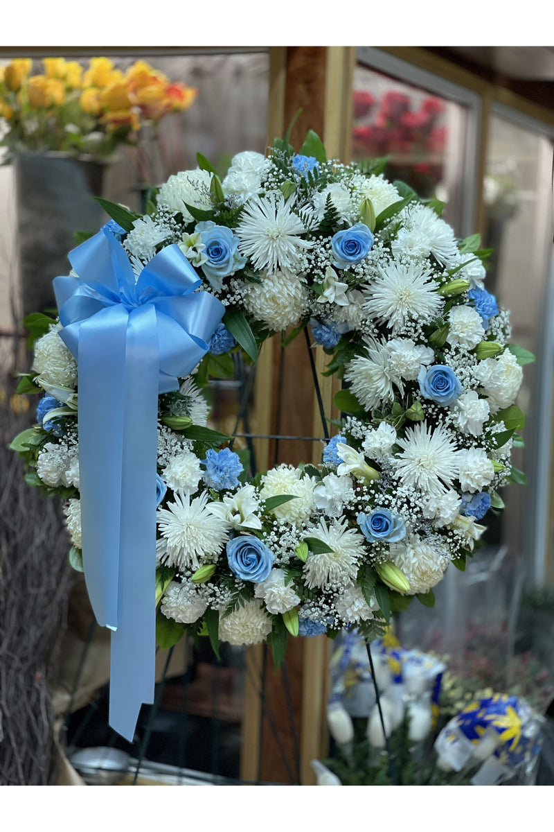 White and blue funeral wreath with white mums, lilies, blue roses, and large blue bow – standing sympathy floral tribute.