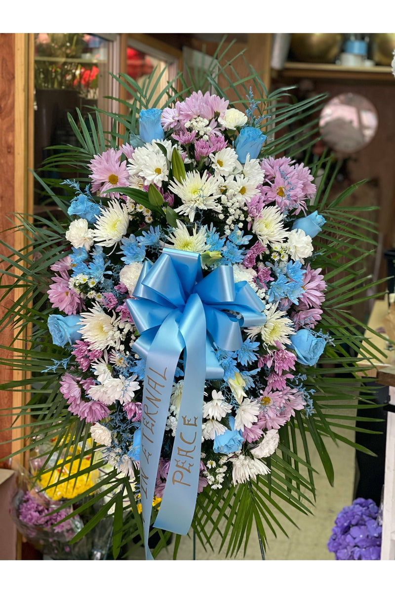 Lavender and blue funeral standing spray with pastel mums, blue roses, white daisies, and blue bow