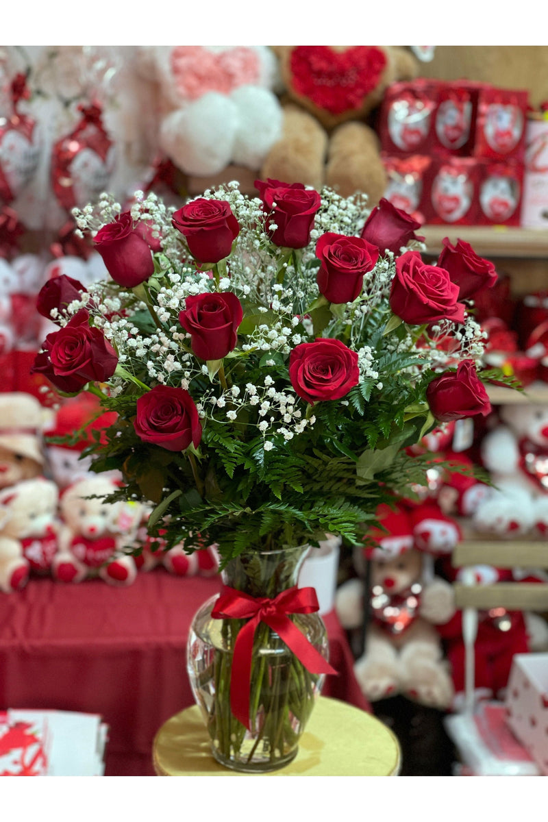 24 red roses with baby's breath and greenery arranged in a glass vase with a red ribbon.