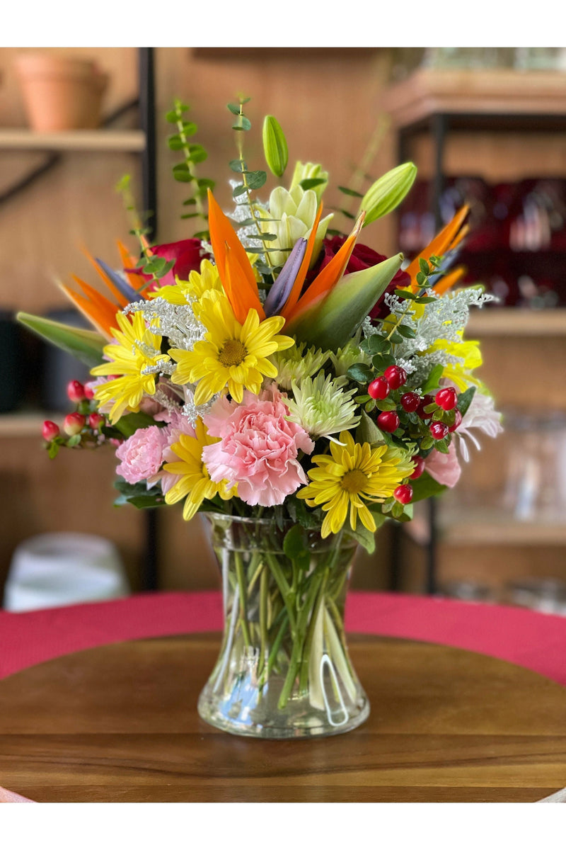 Bright mixed flower arrangement featuring daisies, carnations, birds of paradise, berries, and assorted greenery in a clear vase.