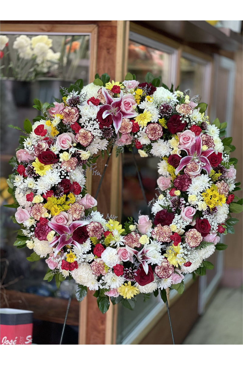 Large funeral tribute wreath with multi-color roses, lilies, carnations, and chrysanthemums arranged in a full circle, handcrafted by Eclore Floral in New Jersey.