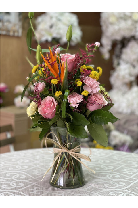 Berry Gum bouquet with pink roses, carnations, Bird of Paradise, yellow poms, and greenery in a glass vase.