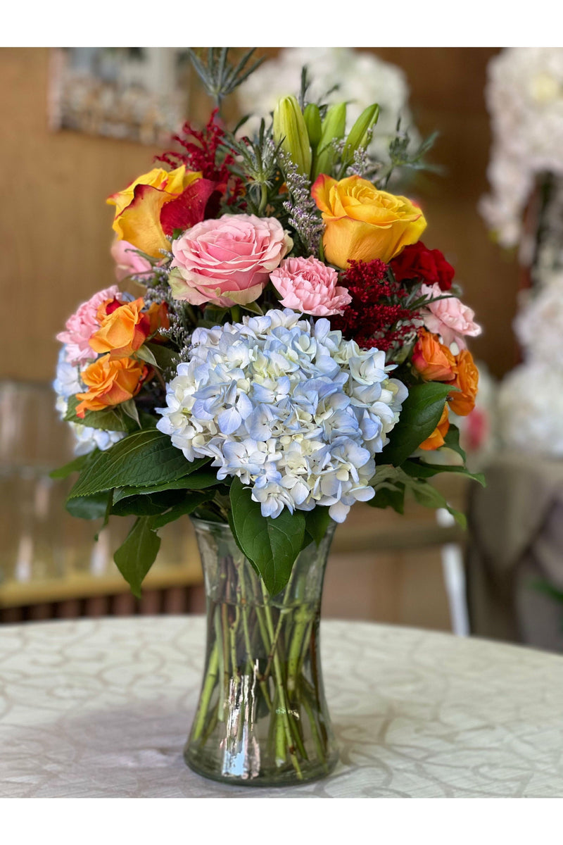 Vibrant tropical flower arrangement with blue hydrangeas, yellow roses, pink roses, and lilies in a clear vase – New Jersey florist.
