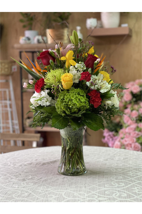 Tropical mixed flower arrangement in a glass vase featuring roses, green hydrangea, Birds of Paradise, lilies, and carnations.