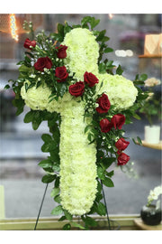 White chrysanthemum funeral cross standing spray decorated with red roses and greenery on an easel.