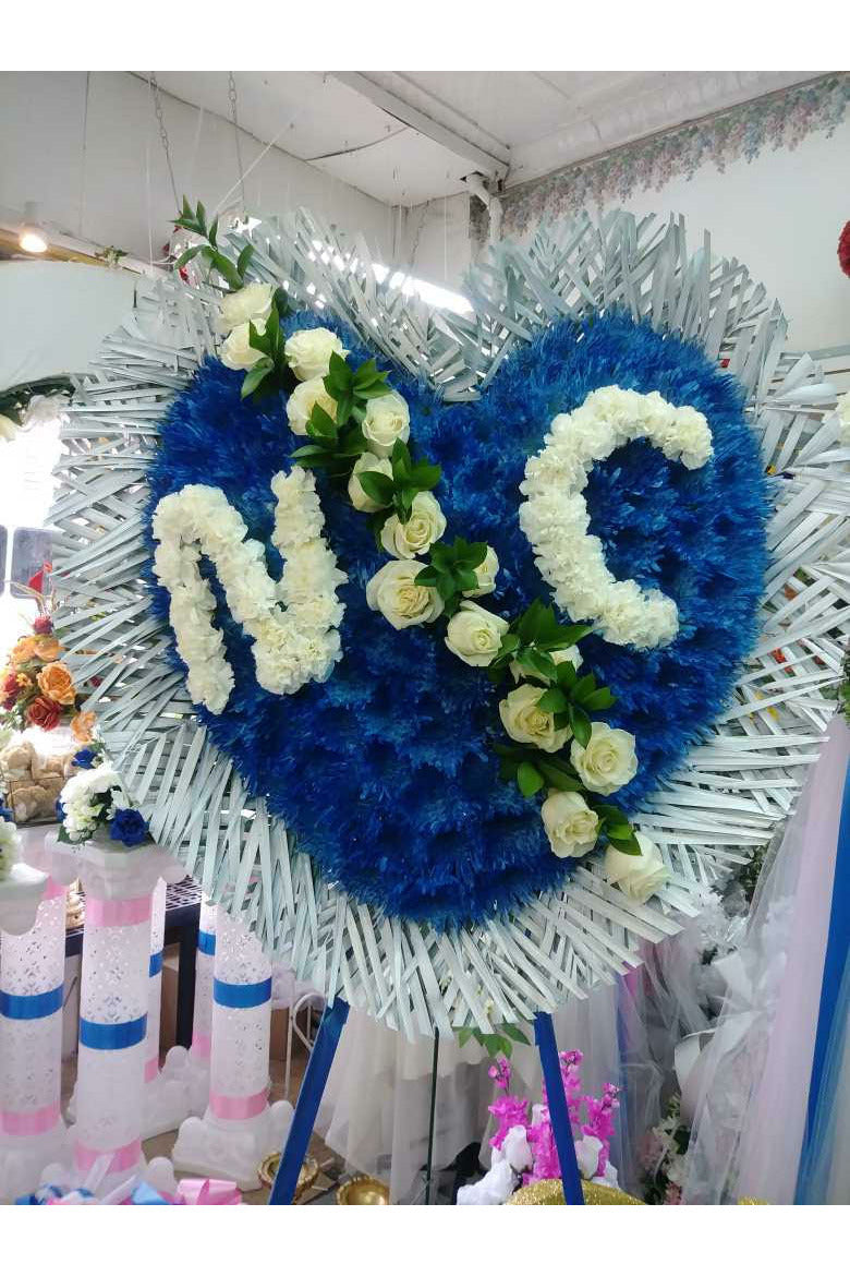 Blue floral heart standing spray with custom white rose initials and greenery – funeral tribute arrangement
